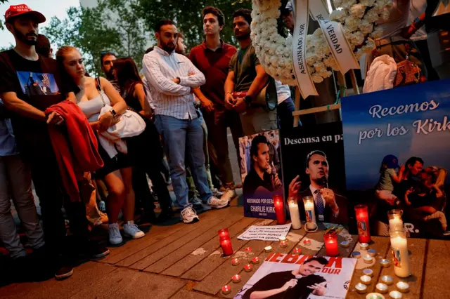 People gather around pictures of Charlie Kirk placed on the ground, next to tea lights and candles. They look down and have sombre faces