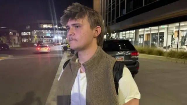 Brock Anderson stands on a street. he is wearing a brown vest and rucksack.