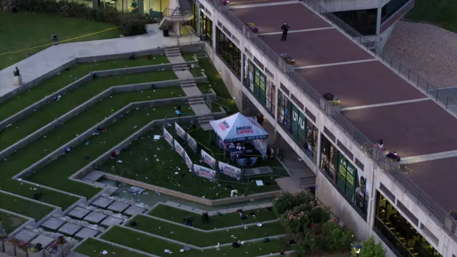 The grassy seating area surrounding the tent is littered with pieces of rubbish. The tent which is branded with the words AMERICAN COMEBACK is still visible. A police officer stands on the roof of the building behind the tent, and police tape cordons off the scene.