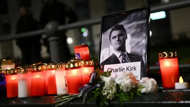 Roses and candles are placed next to a picture of Charlie Kirk during a vigil under the line "In Memory of Charlie Kirk, for freedom, patriotism and justice" in front of the Embassy of the United States