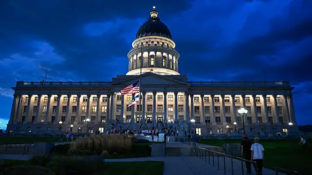 The Utah state capital building in the evening. Several people are gathered outside it and the US flag flies at half mast.