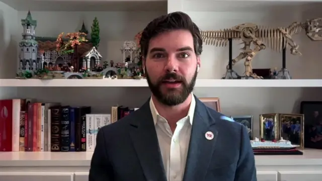 Hayden Padgett wearing a suit speaking in front of a bookshelf, wearing a badge which says 'YR'