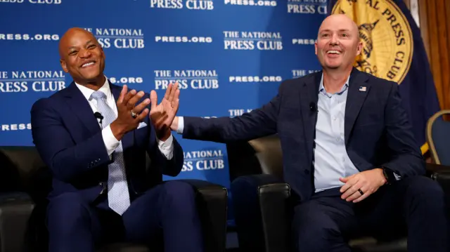 Two bald men in their forties - one black, one white - smile and laugh amiably as they sit on armchairs at a National Press Club event