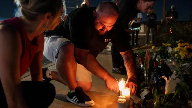 A man wearing a black t-shirt and shorts lights a candle at a vigil for Charlie Kirk.