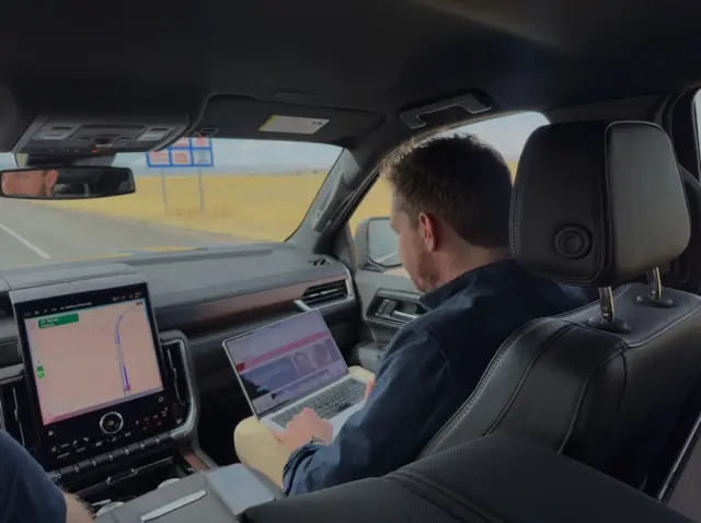 A man sitting in the front seat of a car typing on his laptop