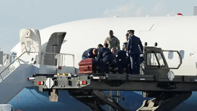 Charlie Kirk's casket is placed on a platform by several security officials. The fuselage of Air Force Two is in the background.