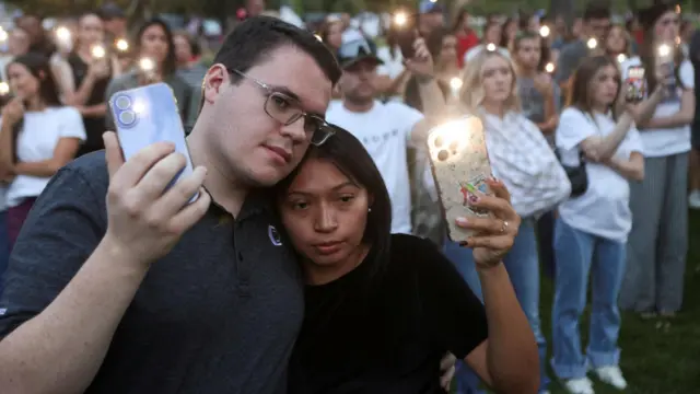 Two people, holding each other, hold up the torches on their phones. Behind them, other attendees also hold up their lit-up phones.