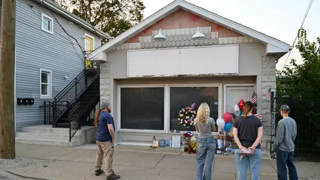 Two men and two women stand outside a one-story house where flowers and US flags have been left in memory of Charlie Kirk. A larger house stands to the left.