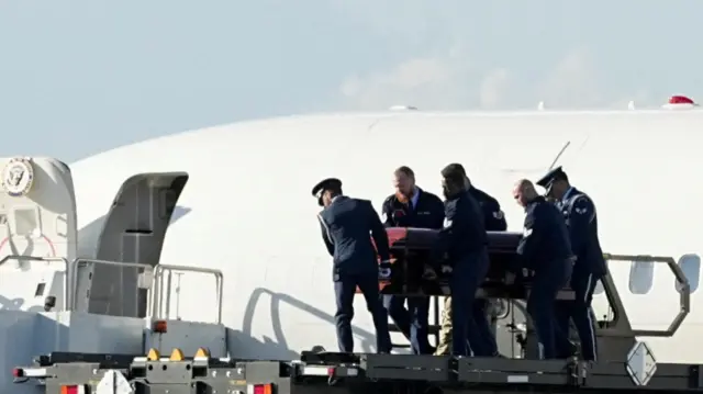 Charlie Kirk's casket is placed on a platform by several security officials. The fuselage of Air Force Two is in the background.