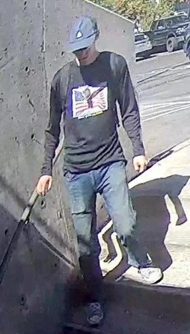 Outside, a person of interest in connection with the murder of Charlie Kirk walks down some stairs. He is wearing a black long sleeve t shit with a white print on the front and a blue blaseball cap