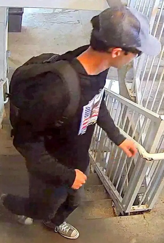 Another image of person of interest in connection with the killing of Charlie Kirk. Walking up a flight of stairs, he is wearing grey All Star trainers, black jeans and a black long sleeve t shirt witha white print of the front. He is carrying a black rucksack and is wearing a baseball cap