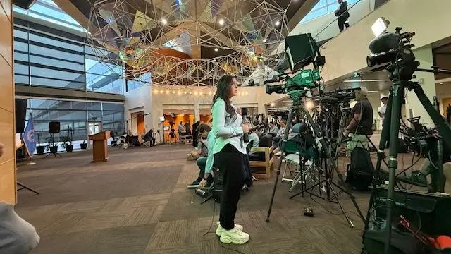 A female reporter stands in front of a line of cameras, in a room with a lone podium in the background.