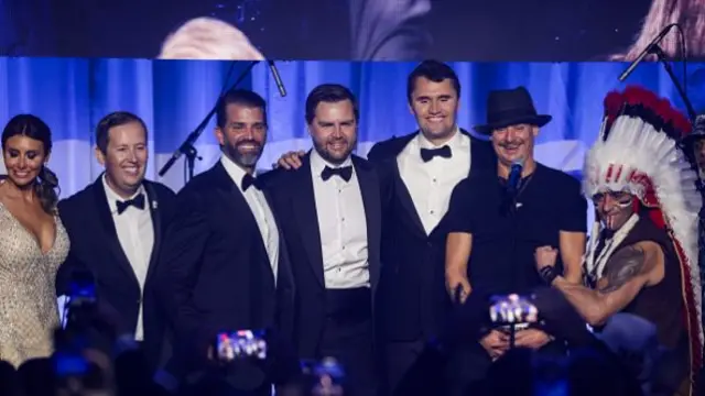Seven people in a line on a blue lit stage, four wear suits, a woman is in a gold dress, and a man is in a native american head dress