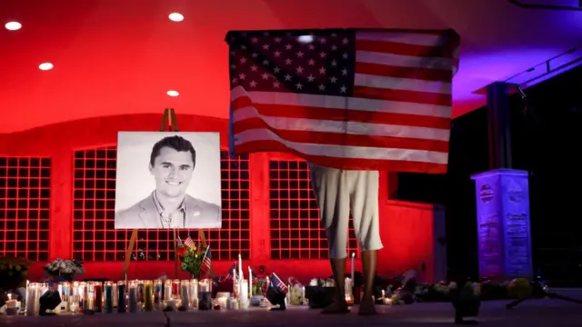 A person holding an American flag stands before a vigil dedicated to Charlie Kirk. A black-and-white photo of the slain activist is on an easel.