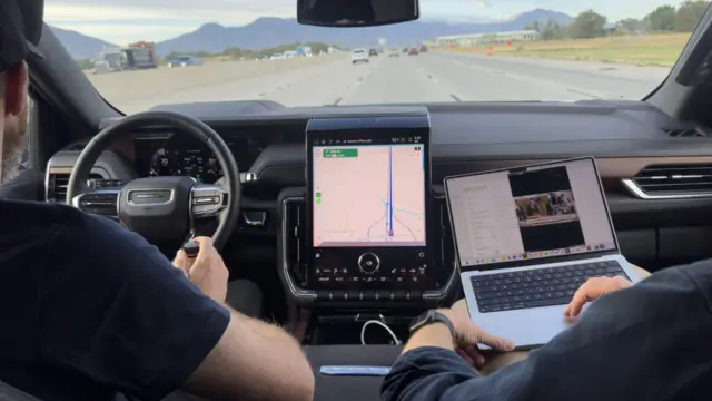 Inside a car on the motorway showing the backs of the driver and someone in the passenger seat working on their laptop, with mountains in the distance