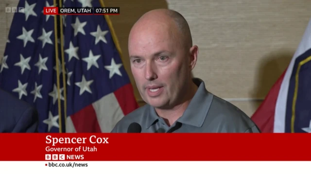 Governor Spencer Cox, wearing a grey shirt, speaks into a microphone at a press conference. An American flag is behind him