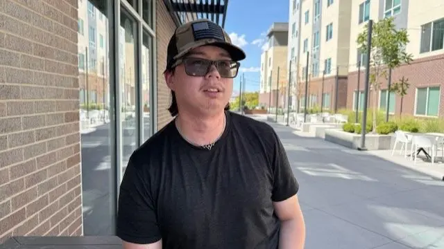 Scott Sperry a young man with light facial hair, sunglasses and a cap, dressed in a black t-shirt, stands in a residential street