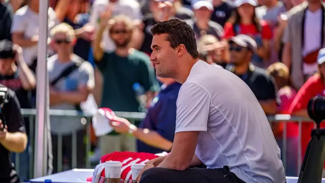 Charlie Kirk sitting down on stage in a white t-shirt moments before his death. A large crowd can be seen standing in the blurred background