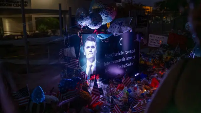 A poster of conservative activist Charlie Kirk is displayed at a memorial, floral tributes, balloons and small US flags around the poster