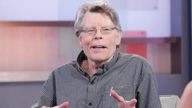 Stephen King in a open-collared shirt mid-speech as he sits on a grey armchair