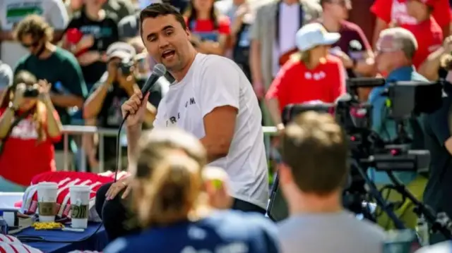 Charlie Kirk in a white t-shirt holding a mic as he speaks while looking to his left. People can be seen standing behind him and in the front of the stage Kirk is sitting on