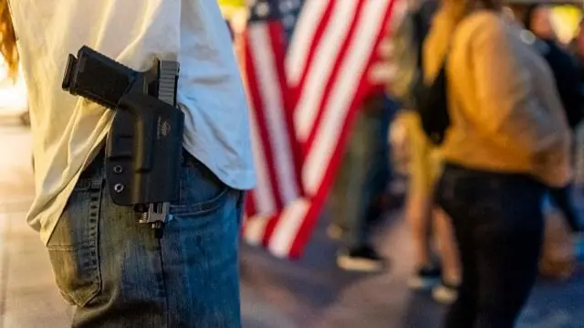 A man in jeans and a grey t-shirt carries a gun in a holster on his hip