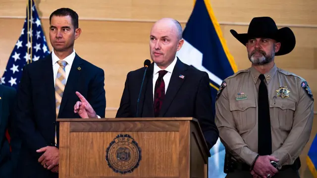 Utah Governor Spencer Cox (centre) stands in a black suit, white shirt and patterned red tie as he speaks at a wooden lectern flanked by another official to his right and the local sheriff to his left