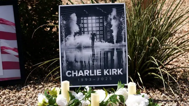 A black and white poster in tribute of Charlie Kirk (showing him walking on a stage, with smoke and sparklers going off around him, the writing Charlie Kirk 1993-2025 at the bottom) lays on the ground. There's three white candles with white flowers placed in front of the poster