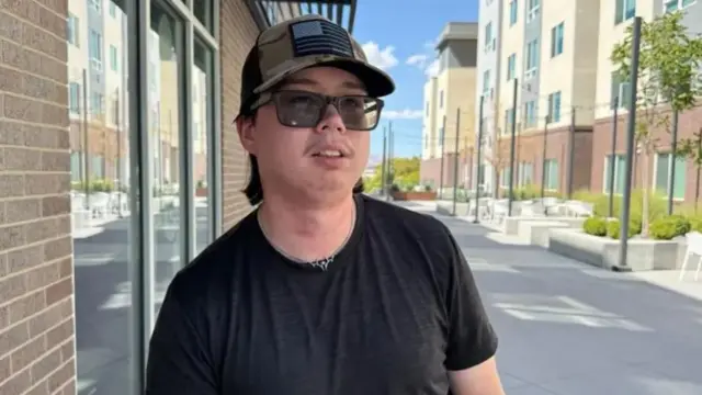 A young man in black shirt, black sunglasses and a military green cap with a a grey-tone US flag on a black background stands next to a brick building, other buildings and greenery to his left