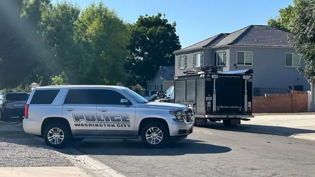A grey Washington City cruiser parked sideways on the street outside a grey two-storey house, a tree-lined street in the background