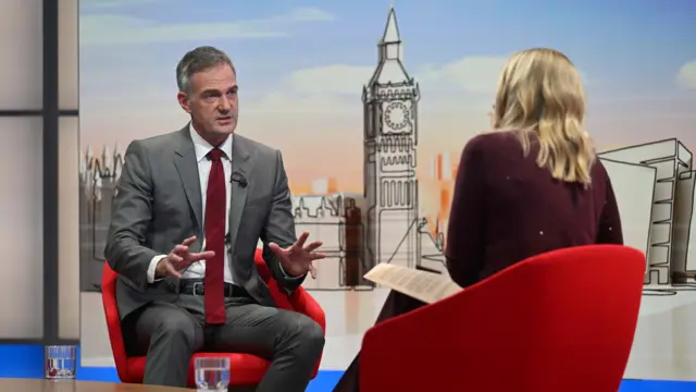 A man in a grey suit and red tie sits in a chair, a blonde woman faces away from him