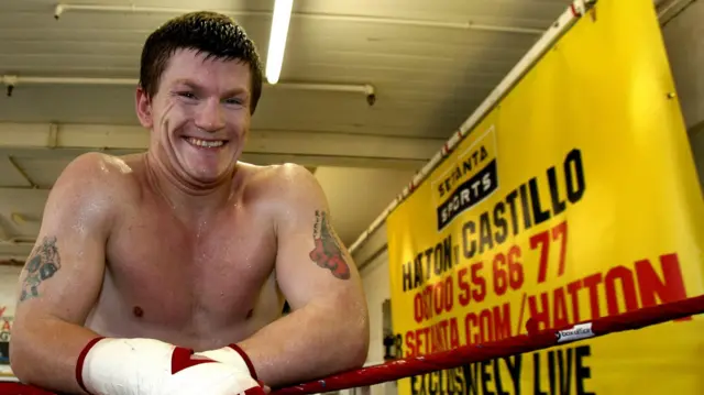 Mid-shot of Ricky Hatton leaning on the top red rope of a ring wearing protective tape around his hands, a yellow fabric sign hanging to his left. The sign reads Hatton v Castillo