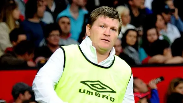 Hatton running on a pitch in front of a stand full of people. He wears a white kit and a bright green Umbro bib
