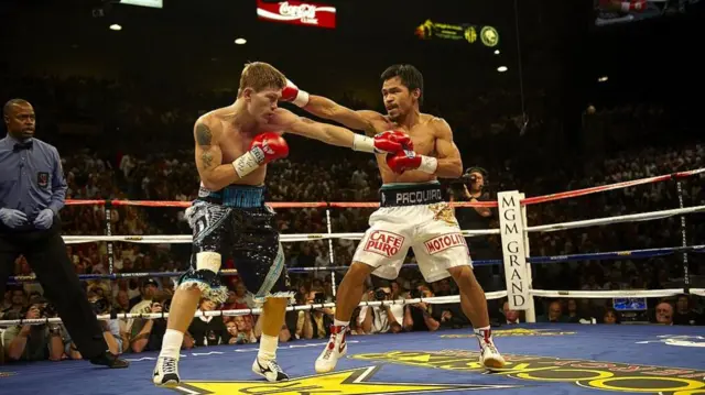 Pacquiao and Hatton in a boxing ring in shorts but shirtless. They wear red boxing gloves and both reach out one arm about to punch each other. There's a huge crowd behind them as well as a camera man