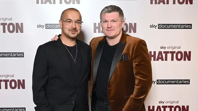 Hatton in a velvet jacket next to a man in a black jacket