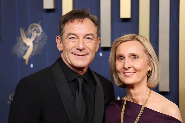 Jason Isaacs, in a black suit, with his wife Emma Hewitt, in a purple dress