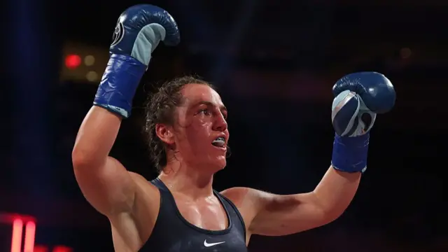 Scotney wears boxing gloves and holds both hands up triumphantly. She is sweaty and wears a sports bra and a gum shield