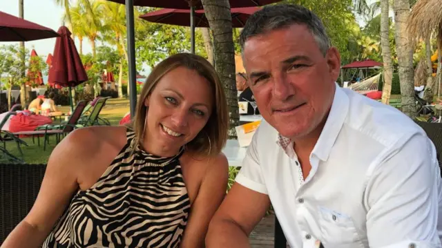 Alex Noble, who has grey hair and is wearing a white button up shirt sitting with his wife Cheryl, who is wearing a zebra-print halter neck dress and has short blonde hair. The pair are smiling at the camera.