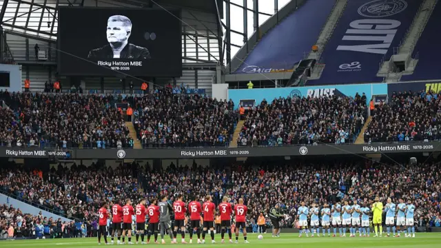 Wide shot of the Eithad stadium in Manchester showing Man United (L in red kit) and Man City (R in blue kit) applaud as a picture of Ricky Hatton is displayed on a large screen on top of the fans