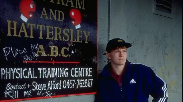 Hatton leaning against a grey wall with has a black sign of a boxing company