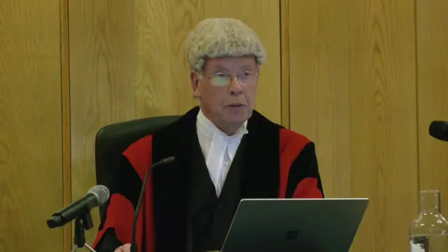 The judge, wearing a robe and wig, speaks.