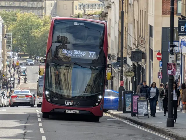 A large red First Bus, number 77 going to Stoke Bishop. It is driving up Park Street in Bristol on a sunny day. There are other cars on the road and lots of people walking on the pavements on either side of the road.