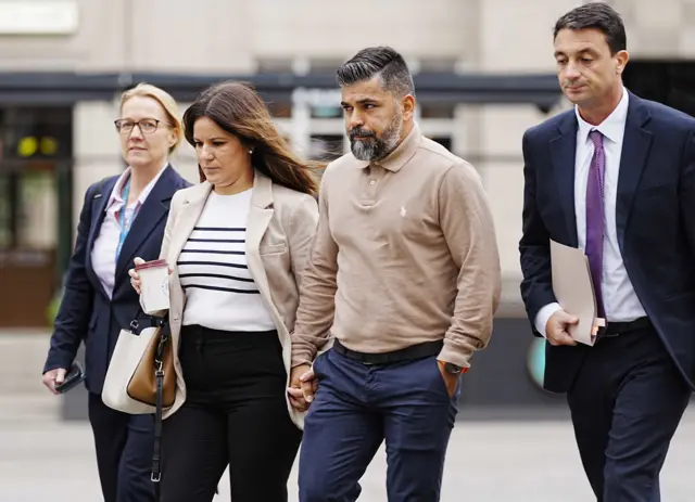 Sergio and Alexandra Aguiar walk across a stone square hand in hand, flanked by legal representatives