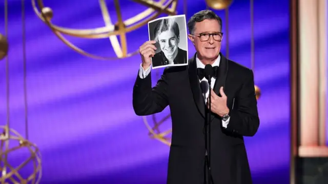 Stephen Colbert holds up a black and white photo of his younger self as he speaks on stage