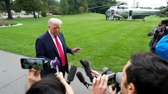 Trump at the centre of the image. He speaks to a group of reporters holding phones and microphones
