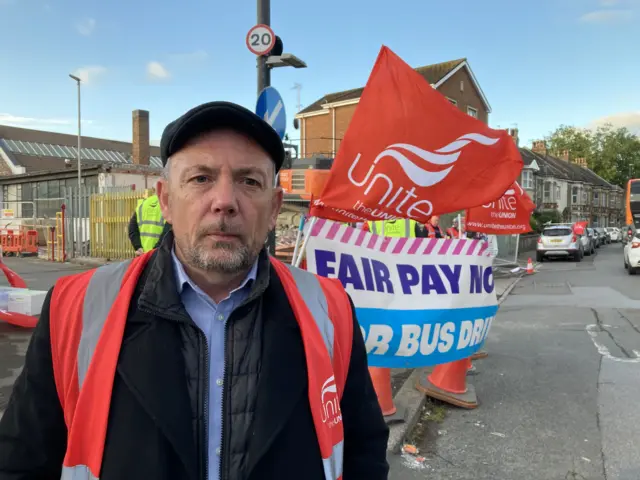 Ken Fish wearing a blue shirt underneath a black jacket and an orange hi-vis vest. He is standing in front of large Unite flags and banners at the picket line.