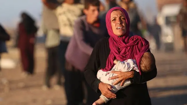 A Palestinian woman wearing a dark pink hijab looks distressed as she carries her small child in her arms.