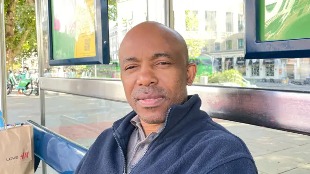 Elo Omyejepu wearing a grey polo shirt underneath a navy fleece jacket. He has a bald head and a closely shaven beard, and is looking at the camera through squinted eyes. He is sitting on a bench at a bus stop in the city centre.
