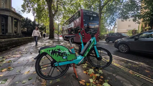 A blue electric bike parked in the foreground. IT has graffiti on the back wheel cover. There is a red bus in the background, and lots of autumnal leaves on the ground.