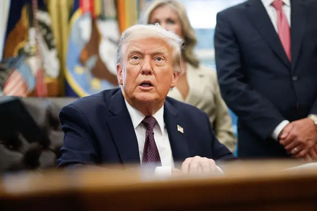 US President Donald Trump speaking in the Oval Office. He is wearig a dark suit and tie with an American flag pin. In the background an aide is visible standing alongside a flag.